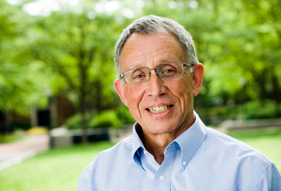 A person wearing glasses and a light blue shirt is smiling in an outdoor setting with trees and greenery in the background.