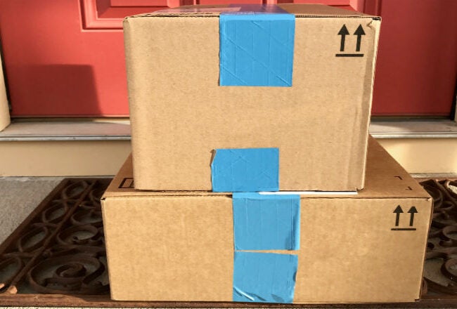 Two cardboard boxes sealed with blue tape are stacked on a doormat in front of a red door.