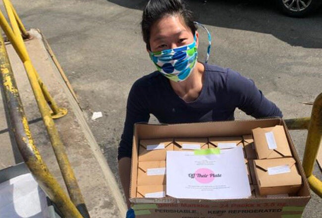 A person wearing a colorful face mask carries a box filled with packaged meals labeled "Off Their Plate" on a sunny day by a stair railing.