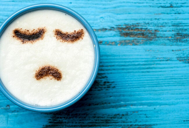 A blue mug with frothy milk on a blue wooden surface. The froth is decorated with cocoa to form a sad face expression.