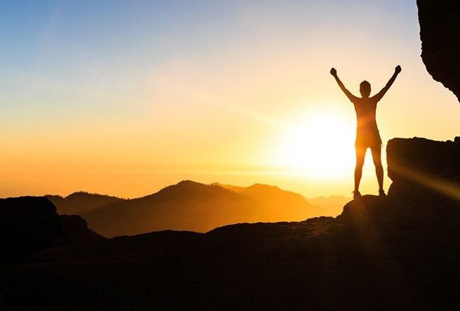 Silhouette of a person with arms raised, standing on a mountain at sunrise, symbolizing achievement and triumph.