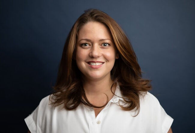 Headshot of a smiling person with long brown hair wearing a white shirt against a dark blue background.