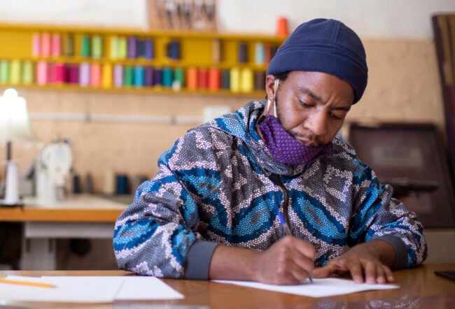 A person wearing a patterned jacket, face mask down, and beanie, writing on paper at a desk with colorful thread spools in the background.