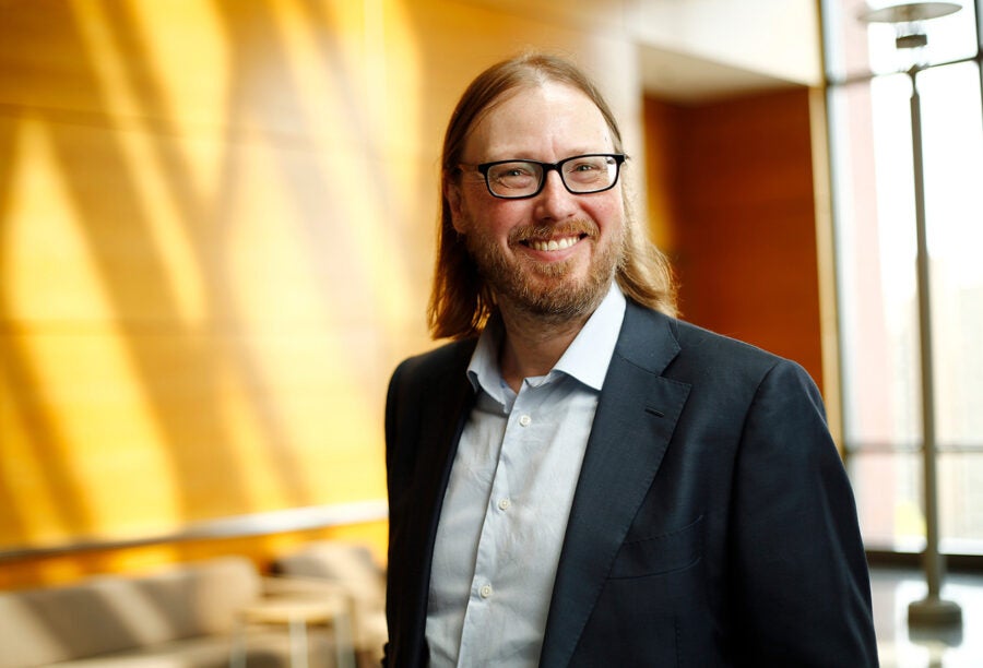 A person with glasses and long hair smiling in a professional setting, wearing a suit and open-collared shirt, with a warm-lit background.