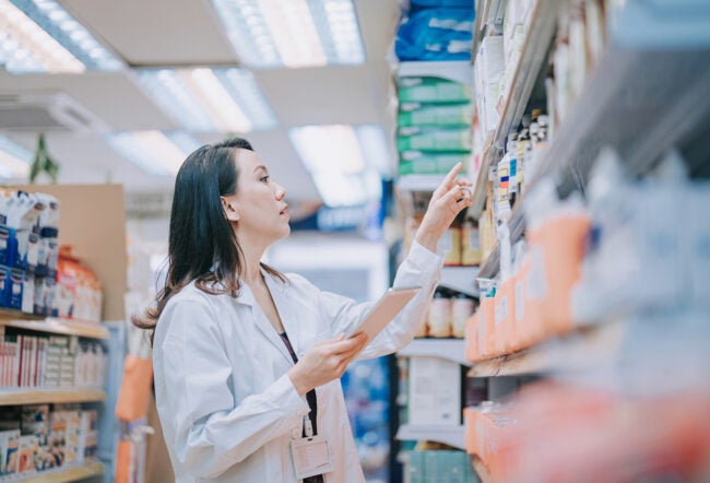 A person in a white lab coat is browsing shelves in a store or pharmacy, holding a tablet and pointing.