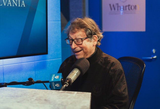 A person with glasses and a dark sweater is speaking into a microphone in a recording studio with blue walls.