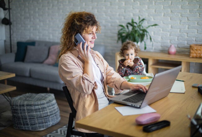 Woman working from home while her toddler is eating in the background, highlighting the importance of addressing caregiver burnout